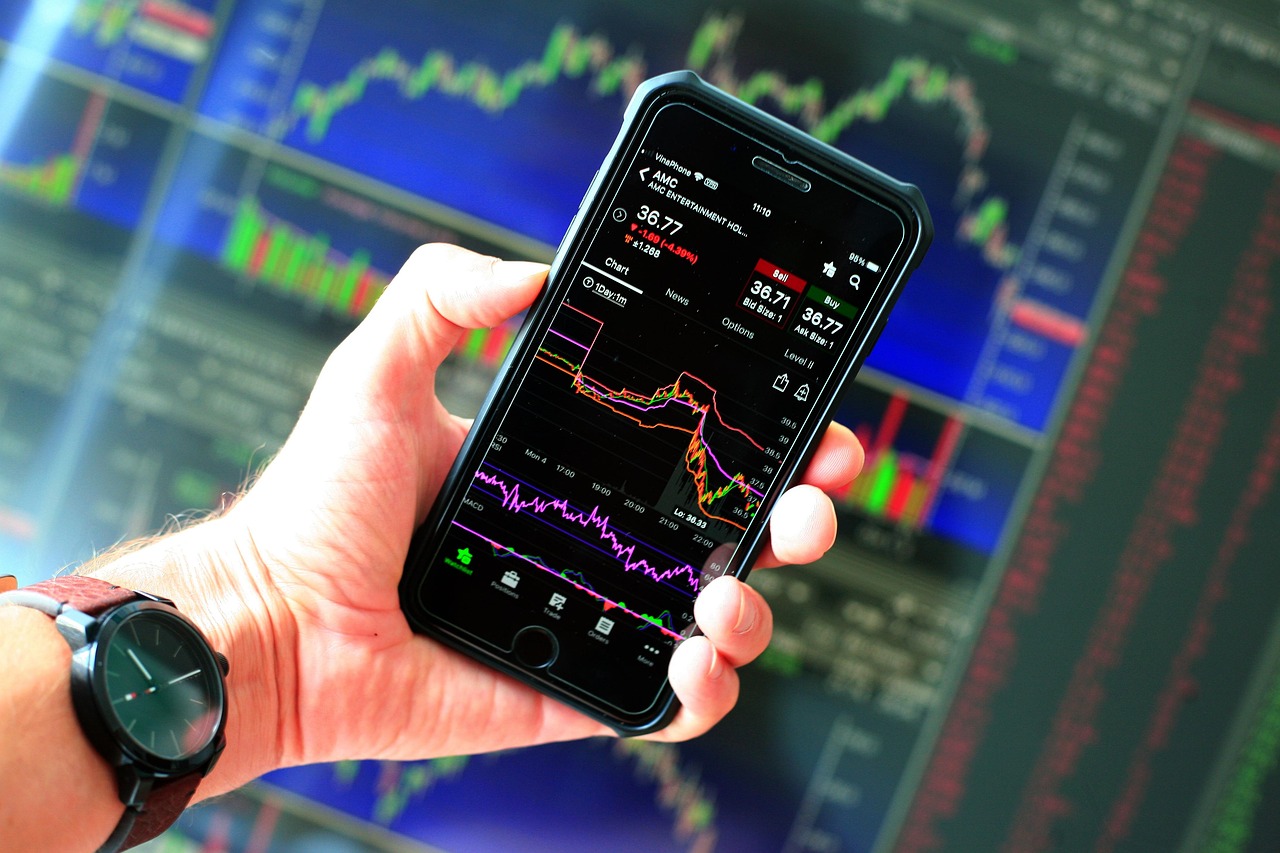 A hand holding a phone showing market data in front of trading screens